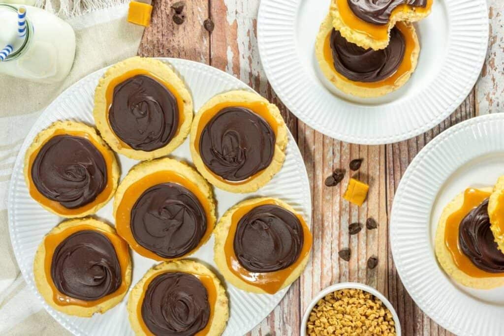 Round cookies topped with a layer of caramel and chocolate are arranged on white plates on a wooden surface, with a small bowl of toffee bits nearby.