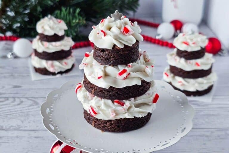 Three stacked brownie rounds with white frosting and crushed peppermint, arranged to resemble Christmas trees, sit on a white cake stand with festive decorations in the background.