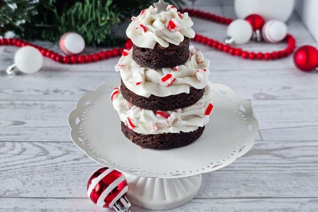 A tiered stack of chocolate brownies with white frosting and crushed peppermint, arranged to look like a Christmas tree, sits on a white cake stand with holiday decorations in the background.