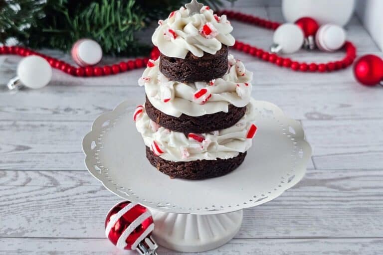 A tiered stack of chocolate brownies with white frosting and crushed peppermint, arranged to look like a Christmas tree, sits on a white cake stand with holiday decorations in the background.