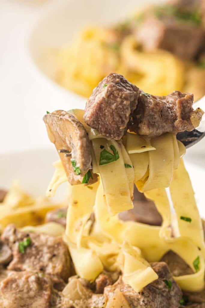 A fork holds up tagliatelle pasta with chunks of beef, mushrooms, and a creamy sauce, garnished with chopped parsley.