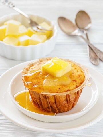 A muffin topped with a pat of melting butter and honey sits on a white plate, with a bowl of butter cubes and two spoons in the background.