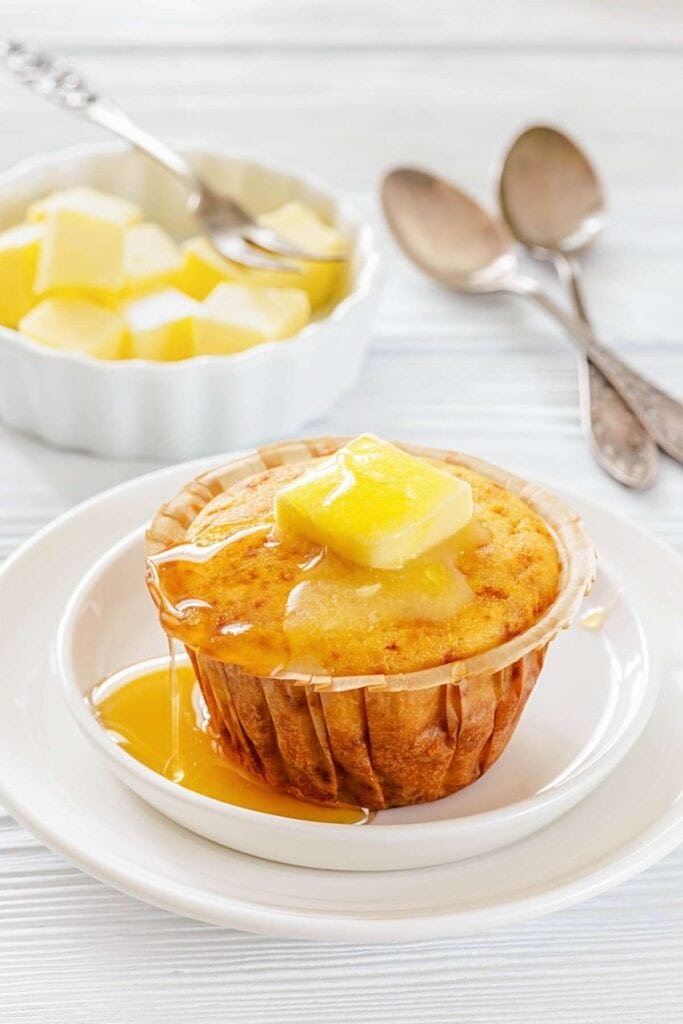 A muffin topped with a pat of melting butter and honey sits on a white plate, with a bowl of butter cubes and two spoons in the background.