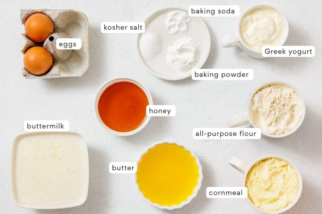 Baking ingredients arranged on a white surface, including eggs, kosher salt, baking soda, Greek yogurt, buttermilk, honey, butter, all-purpose flour, baking powder, and cornmeal.
