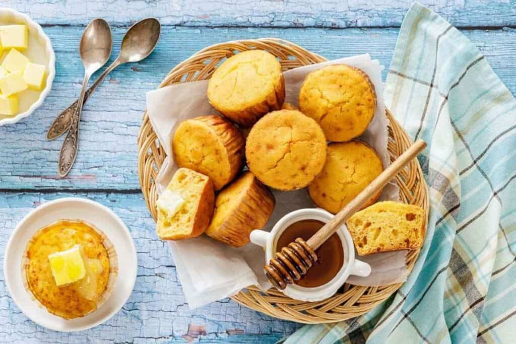 A basket of cornbread muffins sits on a blue wooden surface with honey, a honey dipper, butter, two spoons, and a plaid cloth nearby.