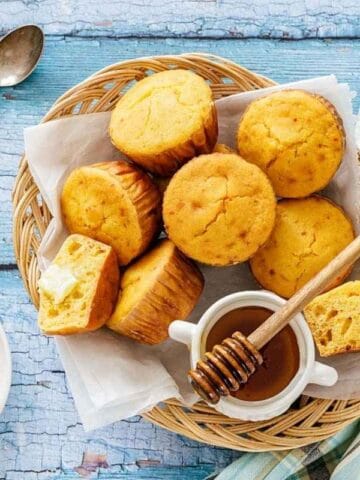 A basket of cornbread muffins sits on a blue wooden surface with honey, a honey dipper, and butter.