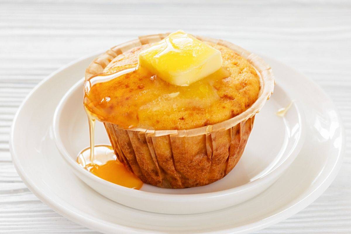 A Honey Cornbread muffin on a white plate, topped with a pat of butter and drizzled with syrup.