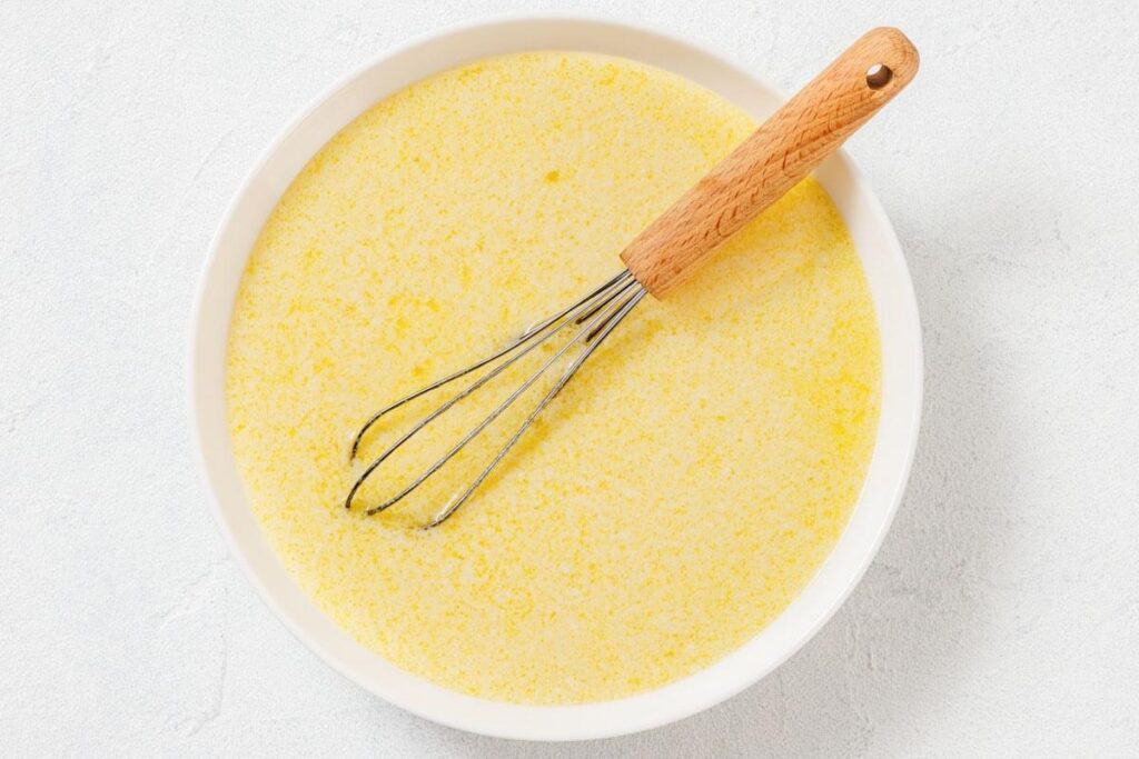 A whisk with a wooden handle rests in a bowl of beaten eggs and milk mixture on a light surface.