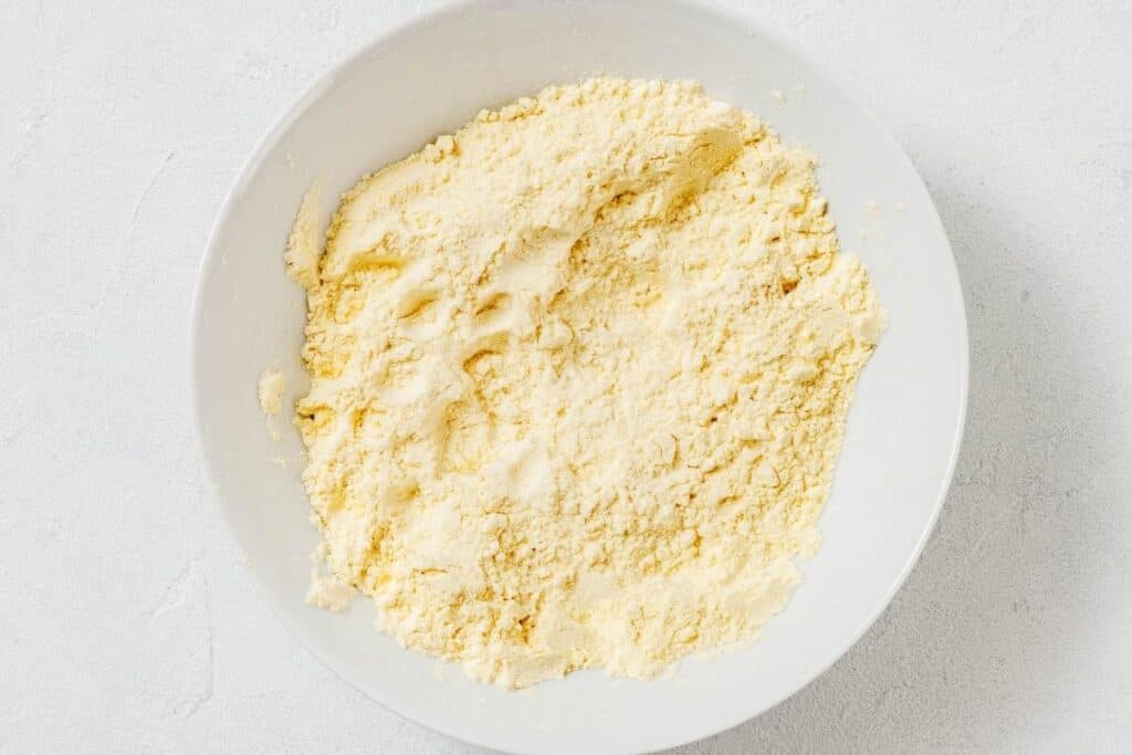 A white bowl filled with a pale yellow, crumbly dry mixture, likely flour blended with butter or another fat, sits on a light-colored surface.
