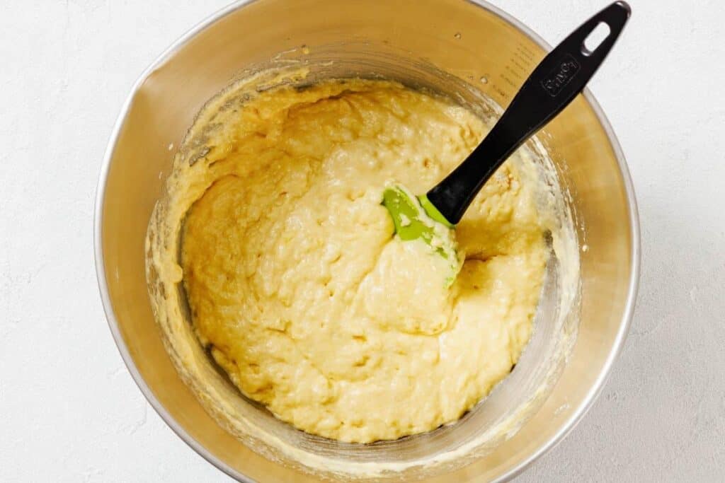 A stainless steel mixing bowl with yellowish batter and a green spatula resting inside.