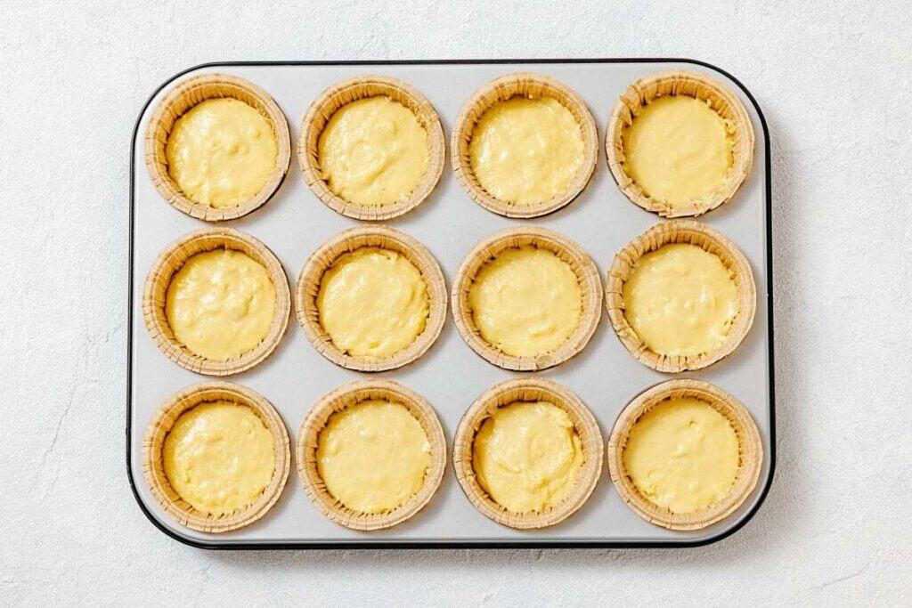 A muffin tray holds twelve unbaked tart shells filled with yellow custard, arranged in three rows of four on a light-colored surface.