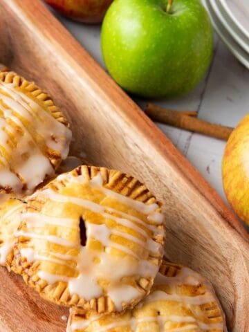 Four glazed apple hand pies are arranged on a wooden tray, with whole apples and stacked plates in the background.