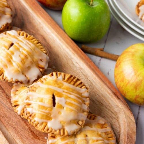 Four glazed apple hand pies are arranged on a wooden tray, with whole apples and stacked plates in the background.