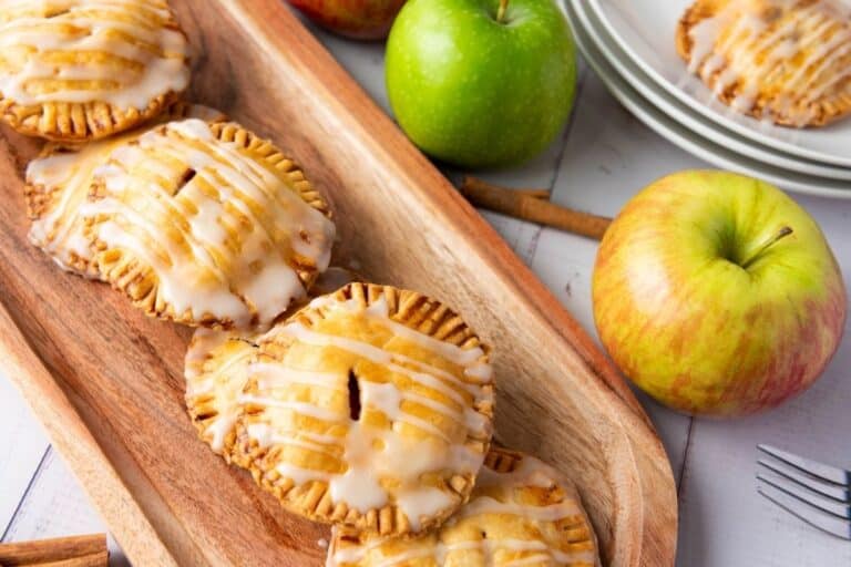 Four glazed apple hand pies are arranged on a wooden tray, with whole apples and stacked plates in the background.