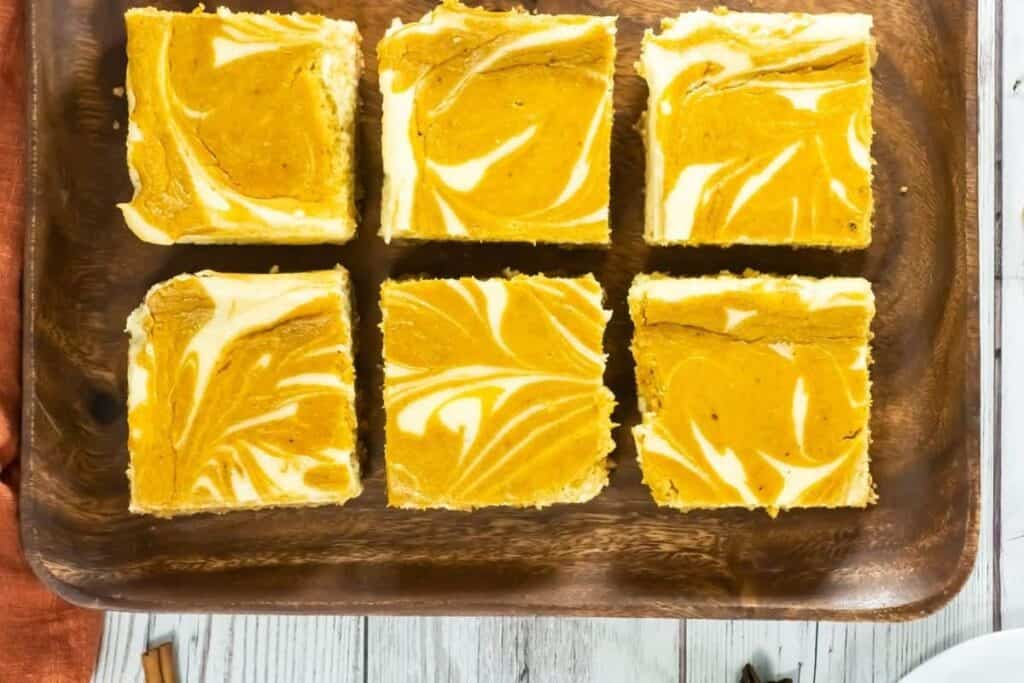 Six square pumpkin swirl cheesecake bars are arranged neatly on a wooden tray, featuring a marbled pattern of orange and white on top.