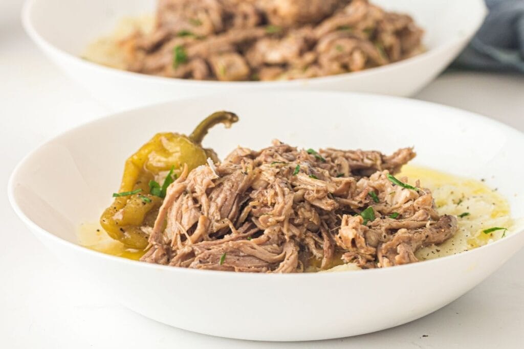 A white bowl filled with shredded beef, mashed potatoes, a pepperoncini pepper, and garnished with chopped herbs.