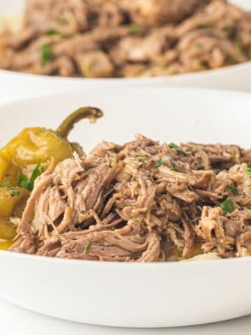 A white bowl filled with shredded beef, mashed potatoes, a pepperoncini pepper, and garnished with chopped herbs.