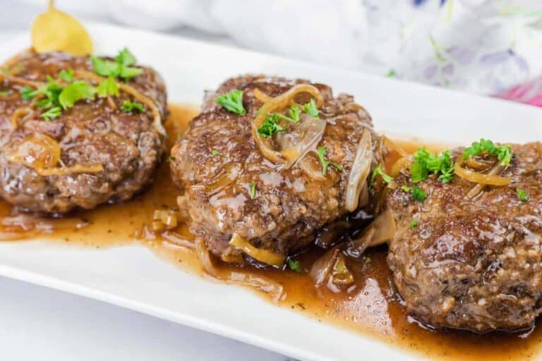 Three hamburger steaks topped with onions and brown gravy, garnished with chopped parsley, served on a white rectangular plate.