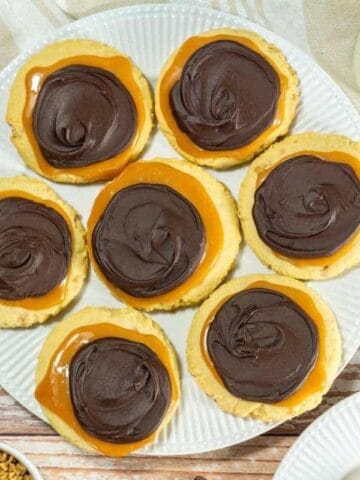 A plate of Crumbl Copycat Twix cookies surrounded by chocolate chips, caramel pieces, a glass of milk, and a beige cloth on a wooden surface.