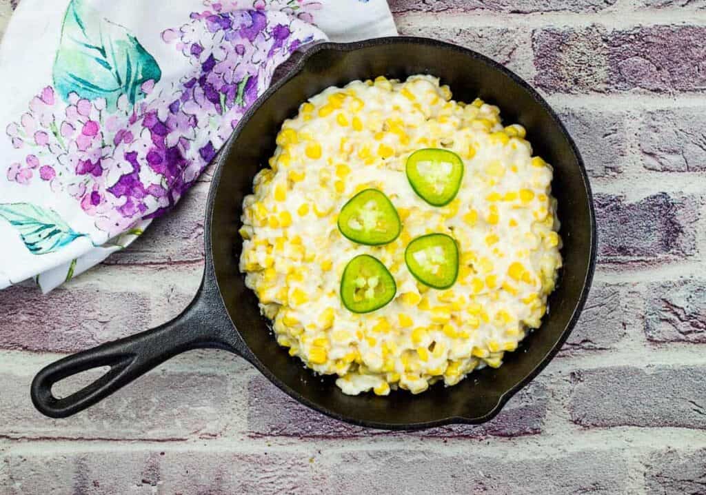 A cast iron skillet filled with creamy corn topped with four jalapeño slices, placed on a brick surface next to a floral-patterned cloth.