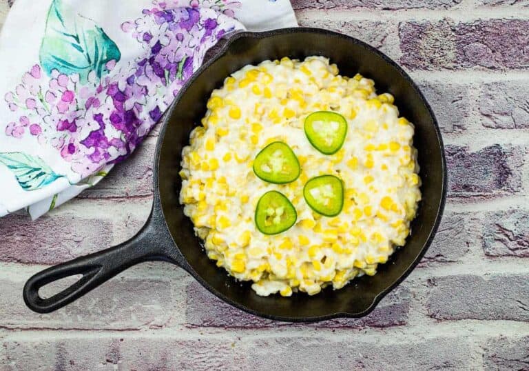 A cast iron skillet filled with creamy corn topped with four jalapeño slices, placed on a brick surface next to a floral-patterned cloth.