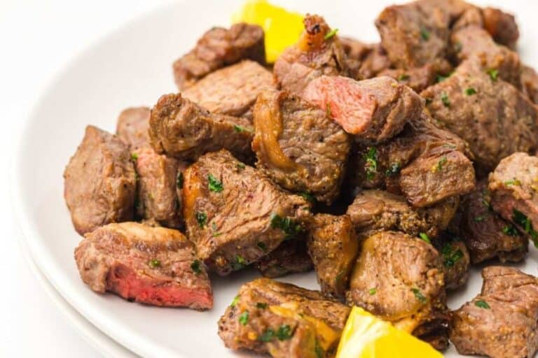 A plate of cooked beef steak bites garnished with herbs, with a few pieces of yellow bell pepper on the side.