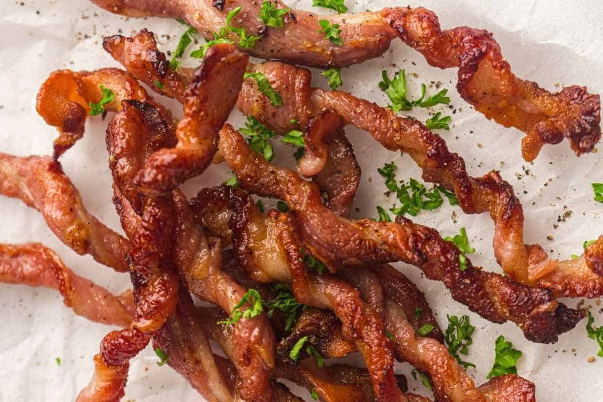 Crispy twisted strips of cooked bacon garnished with chopped parsley, displayed on a white surface.