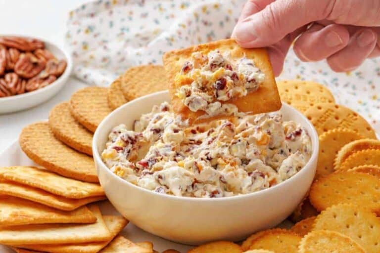 A hand dips a cracker into a bowl of creamy spread with chopped pecans, surrounded by assorted crackers on a white surface.