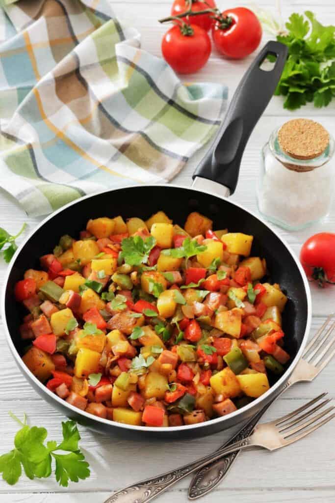 A skillet filled with saut&eacute;ed vegetables including potatoes, carrots, bell peppers, and garnished with fresh herbs, placed on a table with utensils, salt, tomatoes, and a cloth.