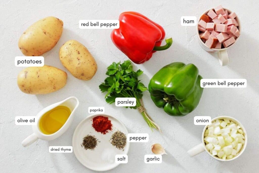 Ingredients for a recipe are laid out on a white surface, including potatoes, red and green bell peppers, parsley, diced ham, olive oil, spices, chopped onion, and a garlic clove.