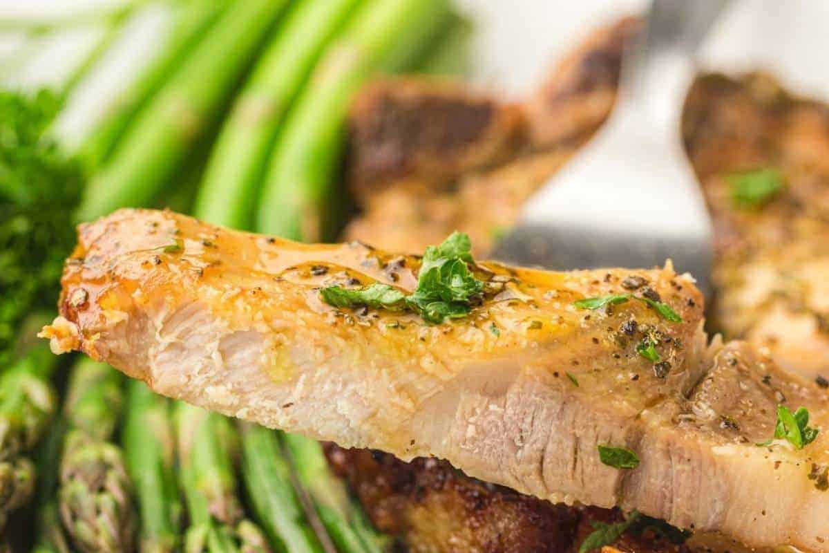 A close-up of a seasoned pork chop slice garnished with herbs, held by a fork, with asparagus spears in the background.