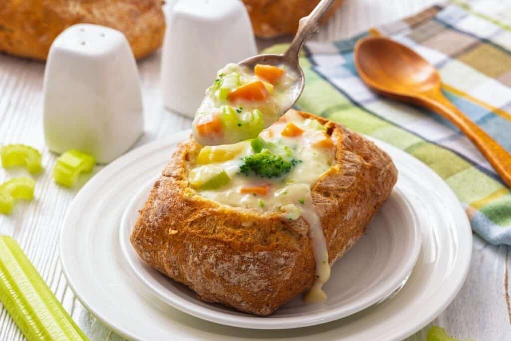 A bread bowl filled with creamy vegetable soup, topped with broccoli and carrots, sits on a white plate with a spoon above it. Celery sticks and salt and pepper shakers are nearby.
