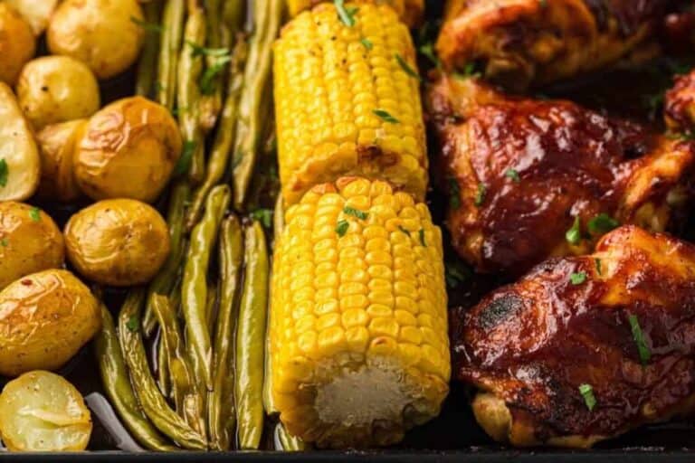 Roasted baby potatoes, green beans, corn on the cob, and barbecue-glazed chicken arranged on a tray.