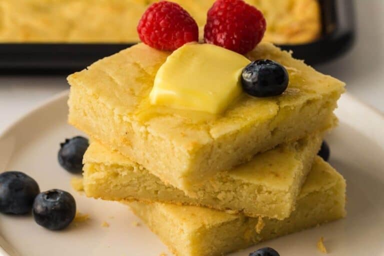 Three pieces of cornbread stacked on a plate, topped with a pat of butter, two raspberries, and a blueberry, with more blueberries scattered around.