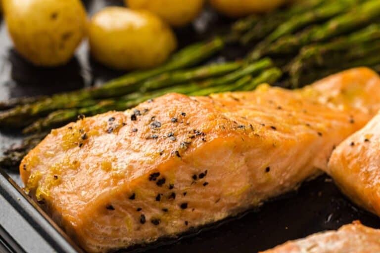 Close-up of baked salmon fillet seasoned with black pepper, with roasted asparagus and potatoes in the background.