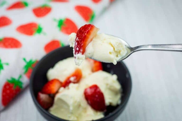 A spoon holding a bite of vanilla ice cream and a strawberry slice, with a bowl of ice cream and strawberries in the background.
