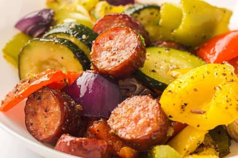 Sliced sausage, zucchini, red onion, and bell peppers seasoned with herbs and spices on a white plate.