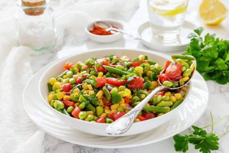 A white bowl filled with a colorful bean salad containing corn, edamame, tomatoes, and green beans, with a spoon resting in the bowl. A glass of water and seasonings are in the background.