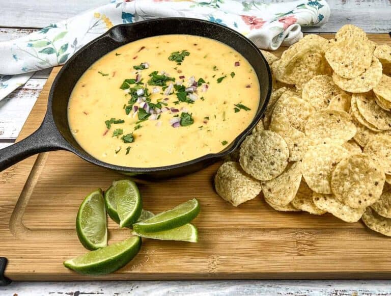 A skillet of queso dip topped with chopped herbs and onions sits on a wooden board next to tortilla chips and lime wedges.