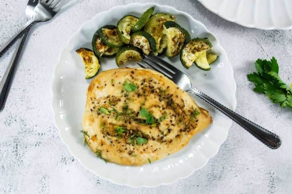A white plate with a cooked chicken breast garnished with herbs and a side of roasted zucchini slices, with a fork resting on the plate.