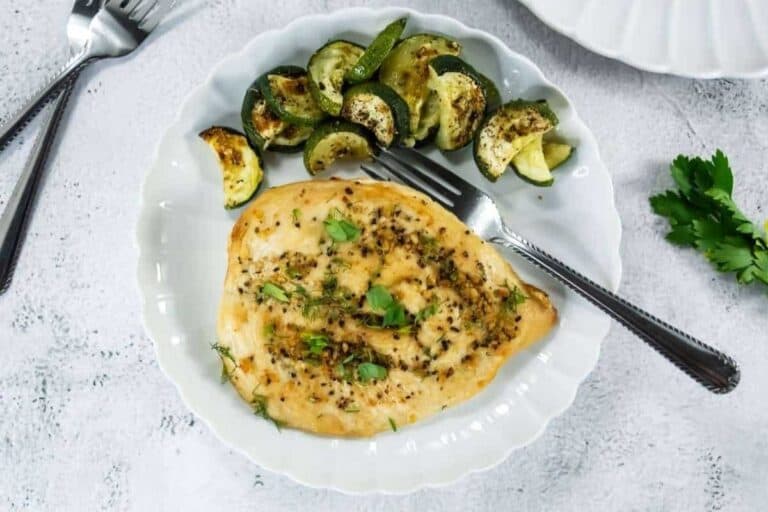 A white plate with a cooked chicken breast garnished with herbs and a side of roasted zucchini slices, with a fork resting on the plate.