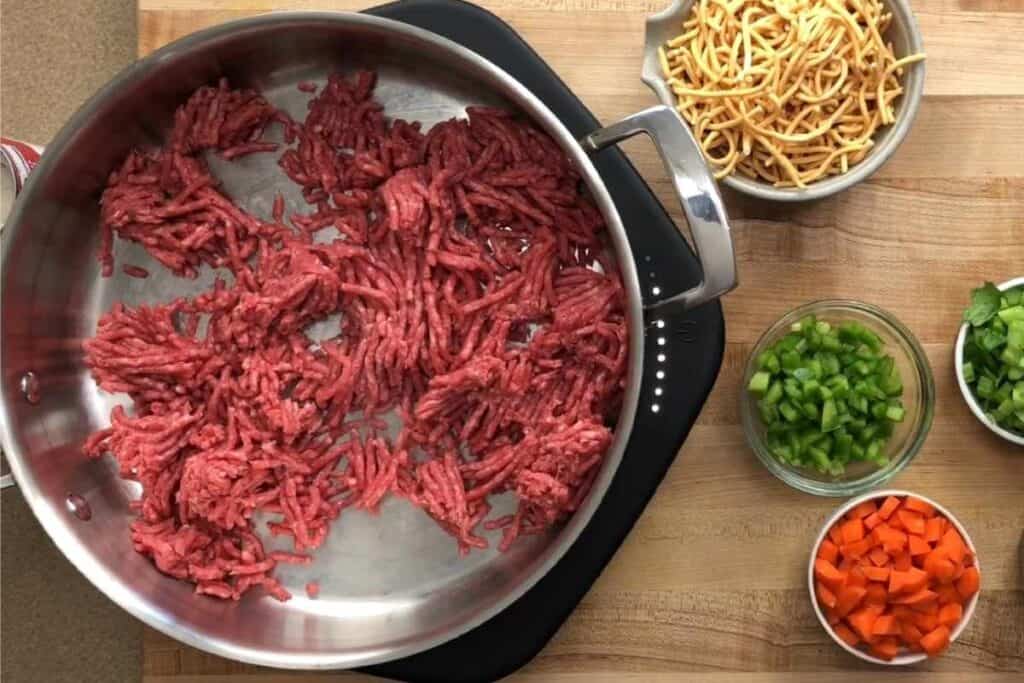 A skillet with raw ground beef sits on a stovetop, surrounded by bowls of chow mein noodles, chopped green pepper, chopped carrot, and chopped celery on a wooden surface.