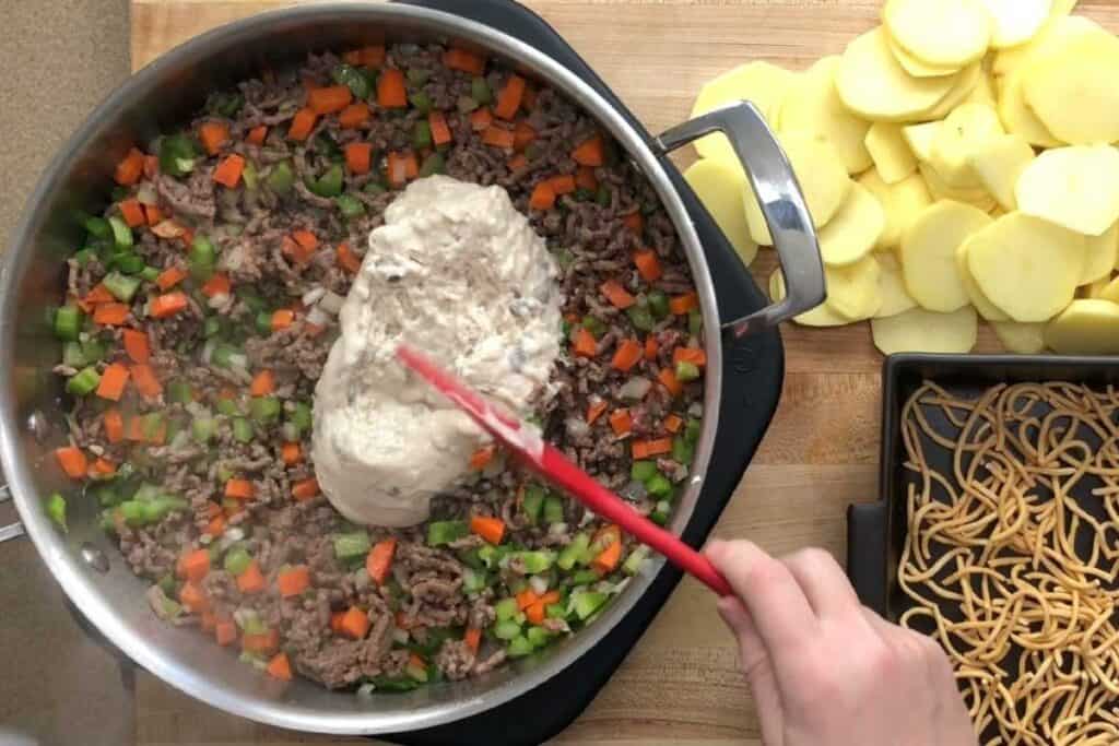 A person stirs ground beef, chopped vegetables, and cream sauce in a pan; sliced potatoes and crispy noodles are on the side.