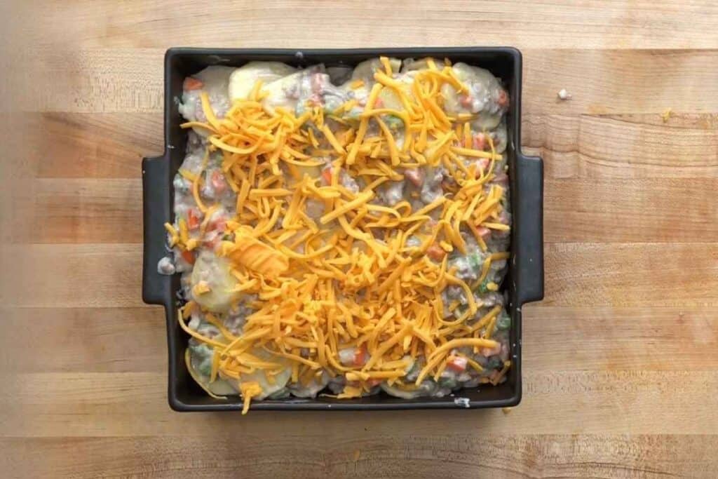 A square baking dish filled with a casserole topped with shredded cheddar cheese, sitting on a wooden surface.