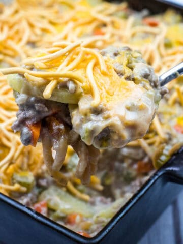 A close-up of a spoon lifting a serving of cheesy casserole with ground beef and vegetables from a square baking dish.