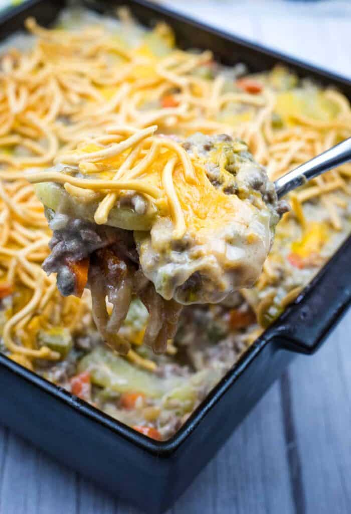 A close-up of a spoon lifting a portion of cheesy casserole with ground beef, vegetables, and shredded cheese from a baking dish.