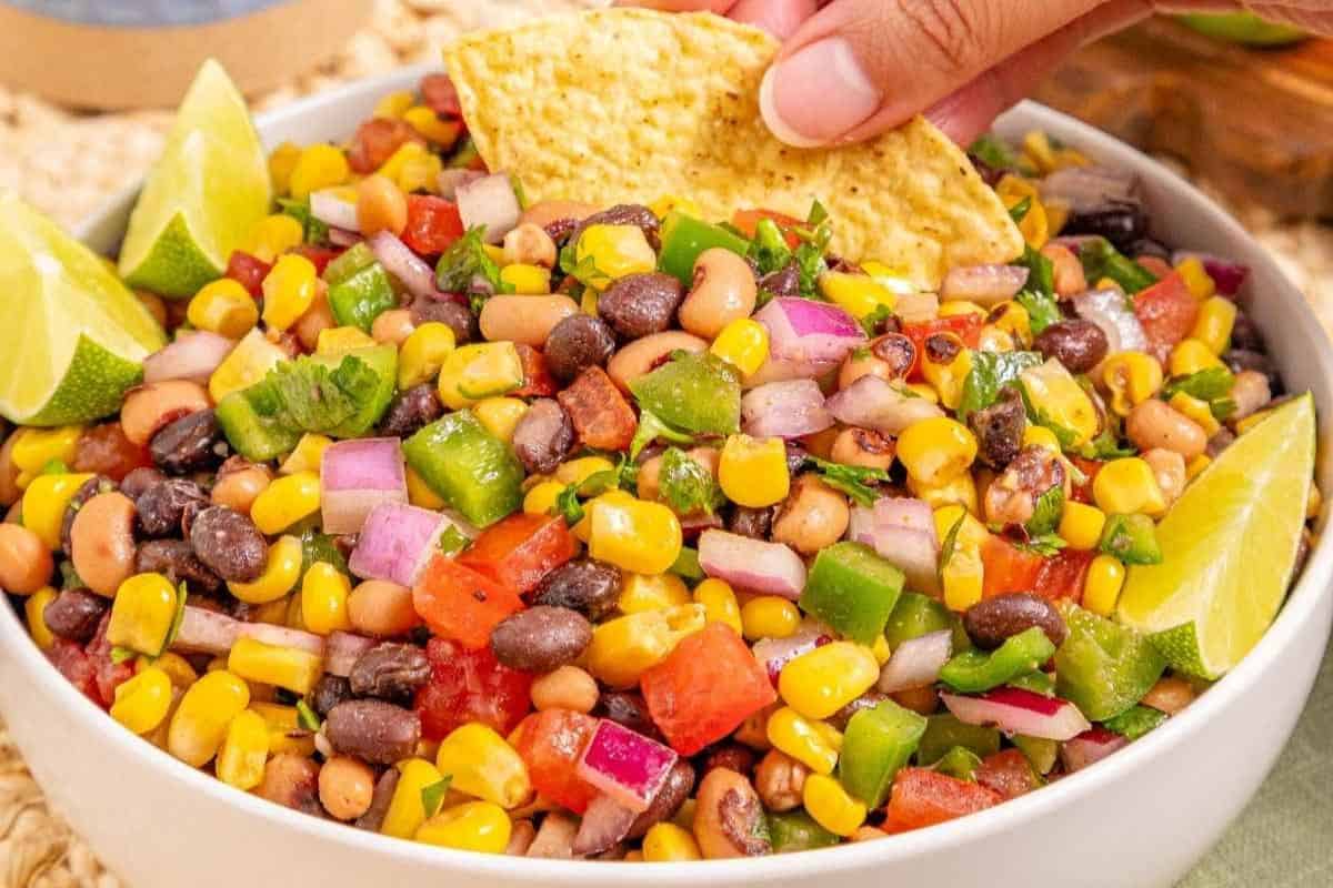 A bowl of Cowboy Caviar, garnished with lime wedges; a hand dips a tortilla chip into the salad.