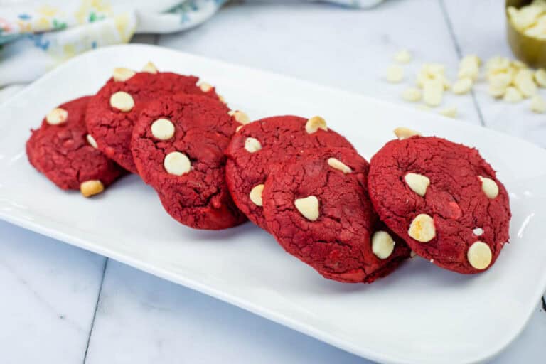Five red velvet cookies with white chocolate chips are arranged in a row on a rectangular white plate, set on a marble surface.
