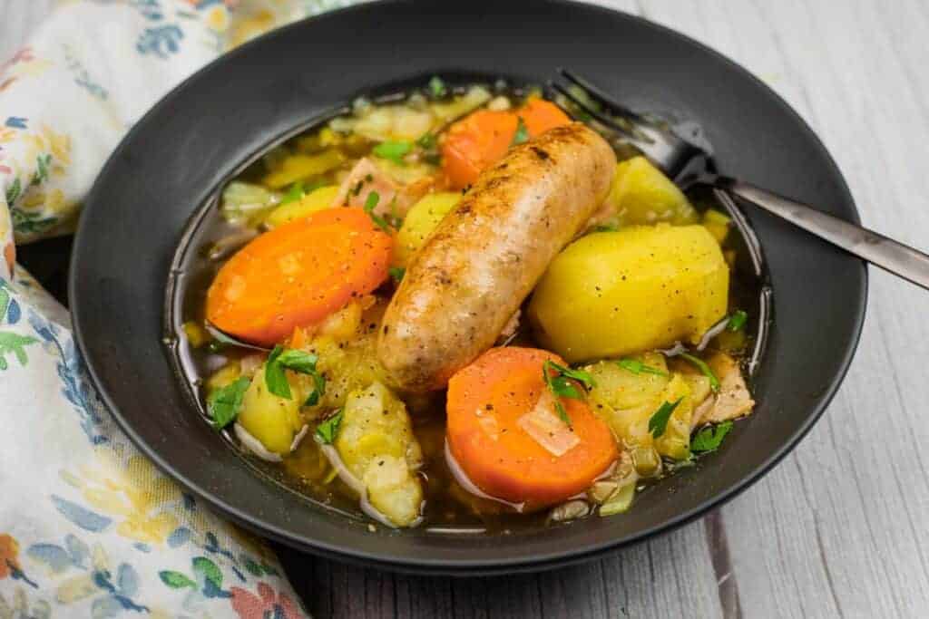 A black bowl of Dublin Coddle filled with sausage, carrots, potatoes, and vegetables in broth, with a fork resting on the edge.