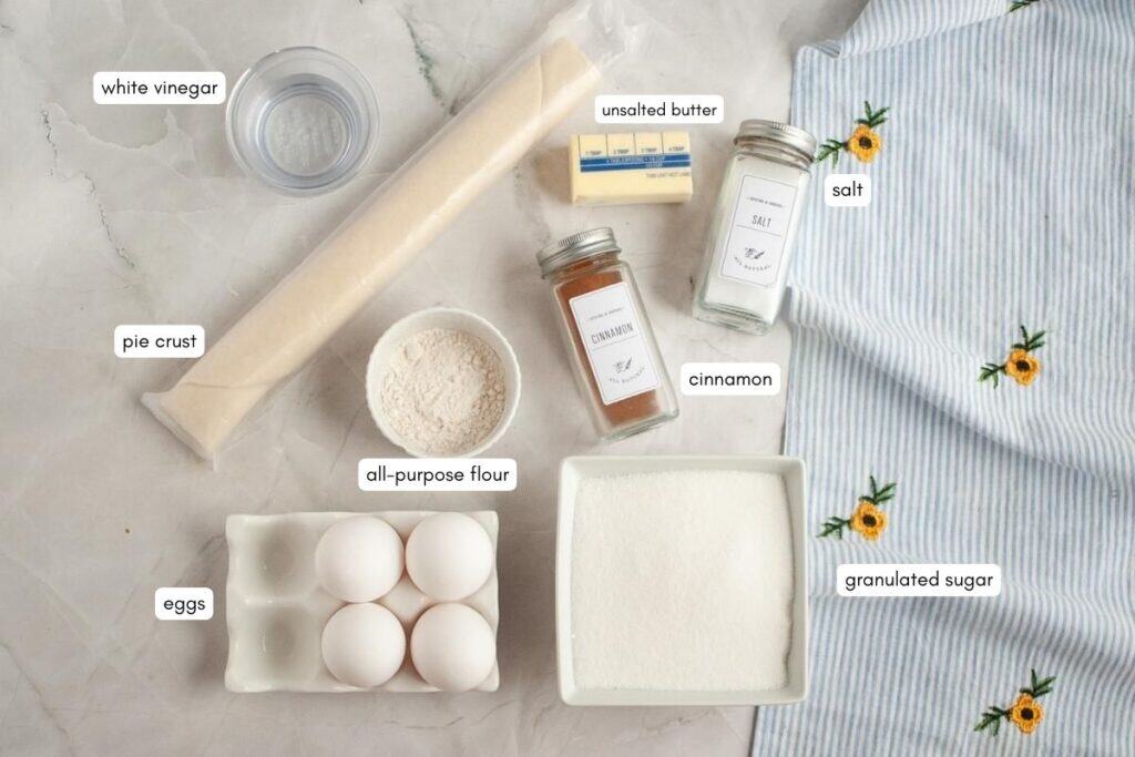 Ingredients for baking are arranged on a white surface, including eggs, sugar, flour, pie crust, cinnamon, unsalted butter, salt, and white vinegar, with labels identifying each item.